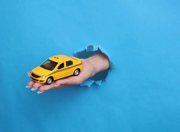 Weibliche Hand hält Spielzeugtaxi-Auto durch zerrissenes Loch aus blauem Papier - Foto, Bild