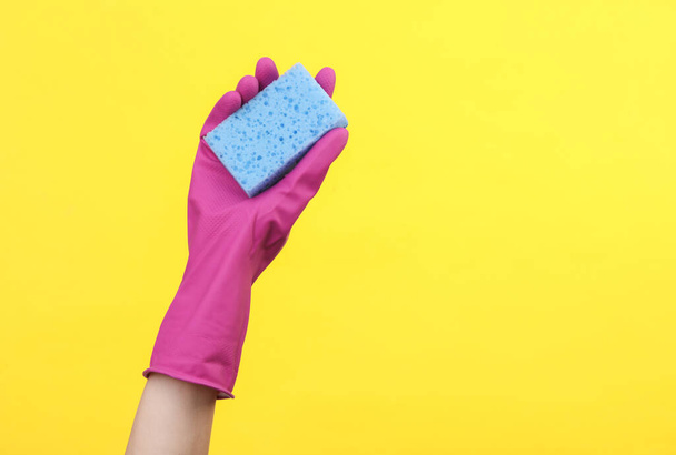 Hand in paars rubber schoonmaak handschoen met spons op een gele achtergrond. Huishouden en schoonmaken concept - Foto, afbeelding