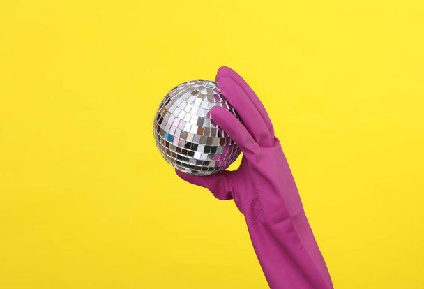 Hand in purple rubber cleaning glove holding disco ball on yellow background. House cleaning and housekeeping concept. Party concept - Фото, изображение