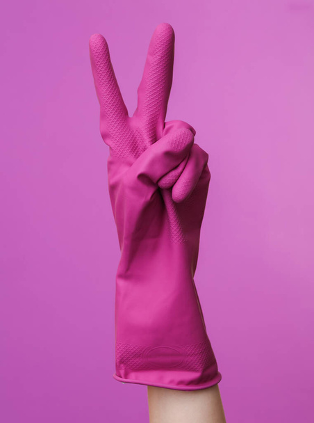 Ręka w fioletowej gumowej rękawicy czyszczącej pokazuje symbol v na fioletowym tle. Koncepcja sprzątania domów i sprzątania - Zdjęcie, obraz