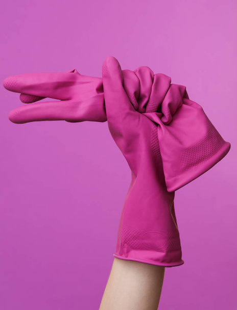 Рука жінки тримає гумові чистячі рукавички на фіолетовому фоні
 - Фото, зображення