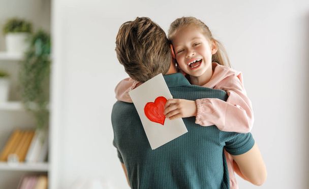 Child daughter is congratulating her father and giving him postcard. Dad and girl are smiling and hugging. Family holiday and togetherness. - Foto, imagen