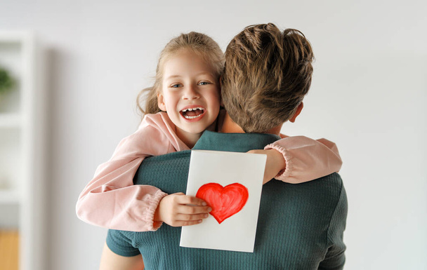 Child daughter is congratulating her father and giving him postcard. Dad and girl are smiling and hugging. Family holiday and togetherness. - Foto, afbeelding