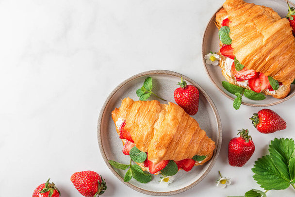 French croissant sandwich with fresh ripe strawberries and cream cheese in a plate with flowers on white background. top view with copy space. tasty breakfast - Photo, Image
