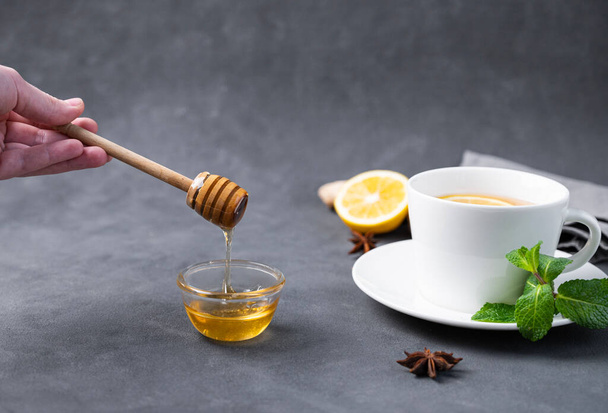 A méz egy fából készült botról egy közeli üvegtálba folyik, sötét háttérrel, egy csésze citrommal és mentával. Az egészséges és biotermékek fogalma. Szabad hely a szövegnek. - Fotó, kép
