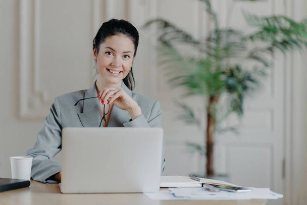 Charming businesswoman freelances on laptop, enjoys remote work, holds glasses, reads publication, sits at desk. - Foto, Imagen