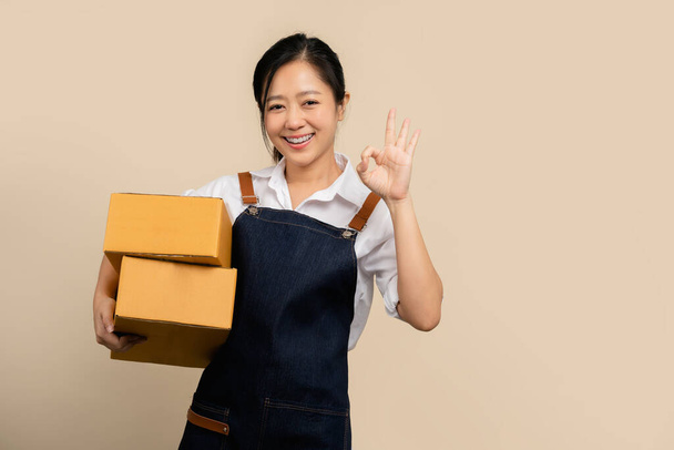 Jovem mulher asiática startup pequeno negócio freelance em avental segurando caixa de pacote isolado sobre fundo marrom claro - Foto, Imagem