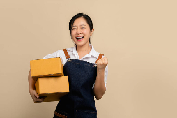 Genç Asyalı bir kadın gülümseyerek küçük bir iş kuruyor ve paket paketini açık kahverengi arka plan, teslimat kuryesi ve nakliye hizmeti kavramına izole edilmiş olarak tutuyor. - Fotoğraf, Görsel