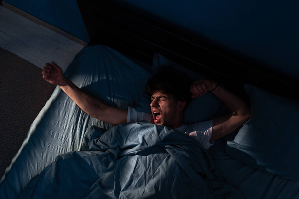 Yawning man awake late at night in bed, can not fall asleep. Sleepy tired, sleep disorder, insomnia and sleeplessness concept. Top view - 写真・画像