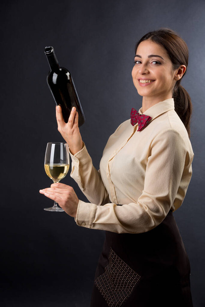 schöne und elegante Kellnerin mit Hemd und roter Fliege ist bereit, ein Glas Wein anzubieten, isoliert auf schwarzem Hintergrund - Foto, Bild