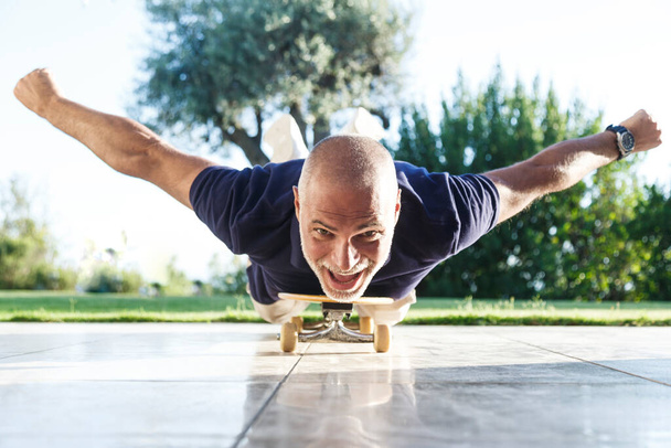Fifty year old man in black shirt and light shorts goes on the skateboard in the prone position - Photo, Image