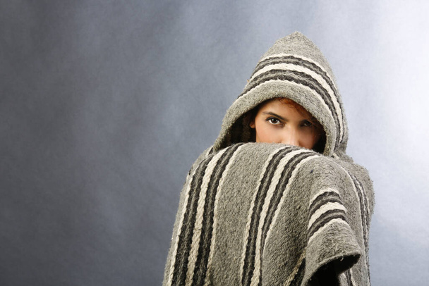 Meisje in poncho verbergt en toont alleen ogen, geïsoleerd op grijze achtergrond - Foto, afbeelding
