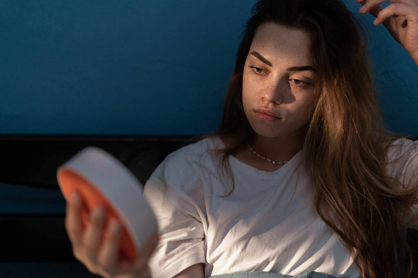 Depressed young woman sitting in bed looking on clock cannot sleep, suffering from insomnia. Sleep apnea, insomnia or stress concept. Copy space - Photo, image