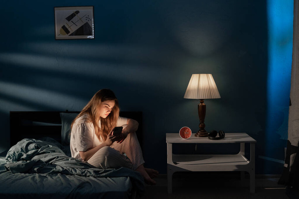 Молодая женщина, сидящая на кровати с помощью мобильного телефона поздно ночью, страдает бессонницей, общаясь в социальных сетях. Концепция интернет-зависимости - Фото, изображение
