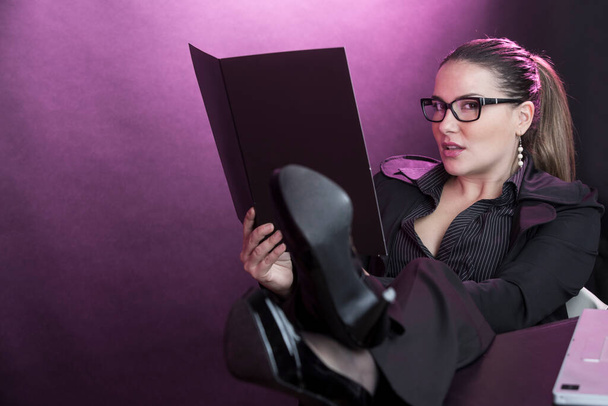 Femme manager avec les cheveux collectés portant des lunettes, habillé en costume d'affaires noir travaillant sur ordinateur avec les pieds sur la table, isolé sur fond rouge - Photo, image