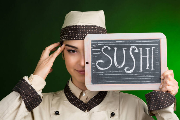 Европейский повар в форме держит доску с суши, написанной на ней, изолированной на зеленом фоне.  - Фото, изображение