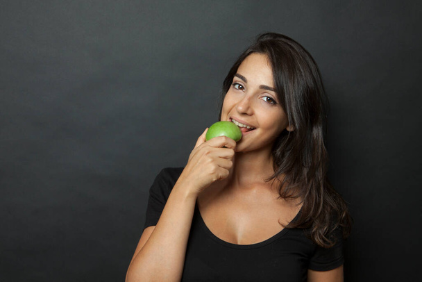 black-haired Woman dressed in black eats an apple happy isolated on black background - Foto, imagen