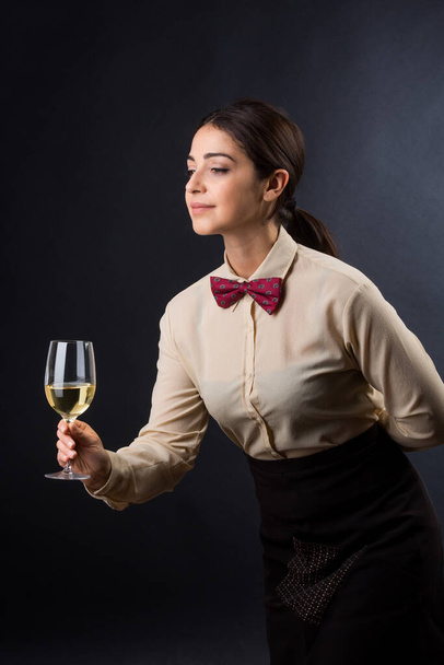 όμορφη και κομψή σερβιτόρα με πουκάμισο και κόκκινο παπιγιόν είναι έτοιμη να προσφέρει ένα ποτήρι κρασί σε έναν πελάτη, απομονωμένη σε μαύρο φόντο - Φωτογραφία, εικόνα