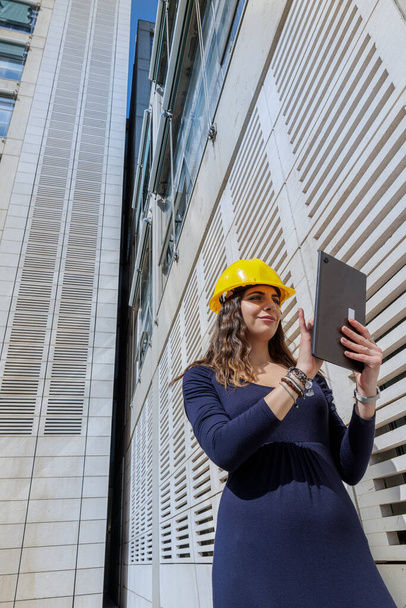 Mujer elegantemente vestida con casco protector amarillo, sosteniendo su tablet en la mano en un contexto urbano moderno - Foto, Imagen