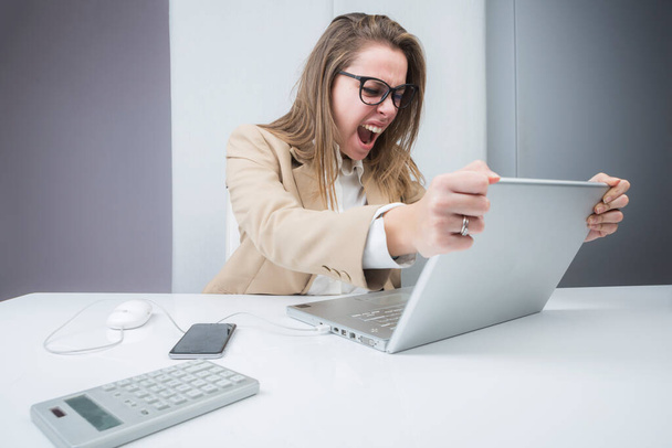 blonde girl dressed in a beige jacket is sitting at her workstation in front of the portal, gets angry and violently shakes the monitor - Photo, Image