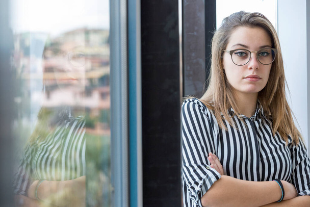 Light-skinned blonde girl in eyeglasses and a striped shirt is standing with her arms folded near the glass window of her office from which other buildings can be seen - Photo, Image