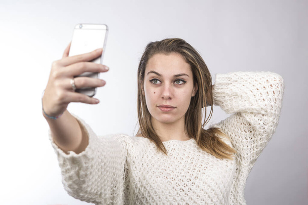 blonde girl in white sweater takes a selfie with her smartphone, isolated on white background - Photo, Image