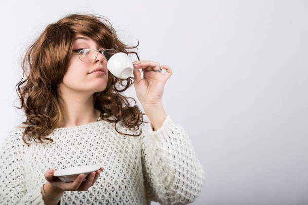 Dame mit braunem welligem Haar und weißem Pullover trinkt ihren Kaffee aus einer Tasse, isoliert auf weißem Hintergrund - Foto, Bild