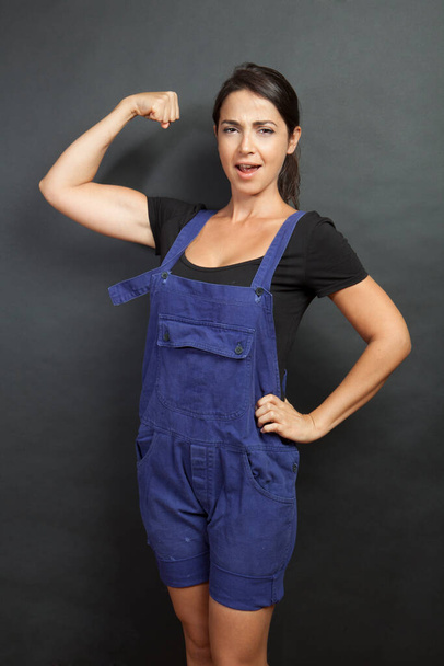 Forced white girl with black hair and work overalls shows the arm muscles, isolated on black background - Valokuva, kuva
