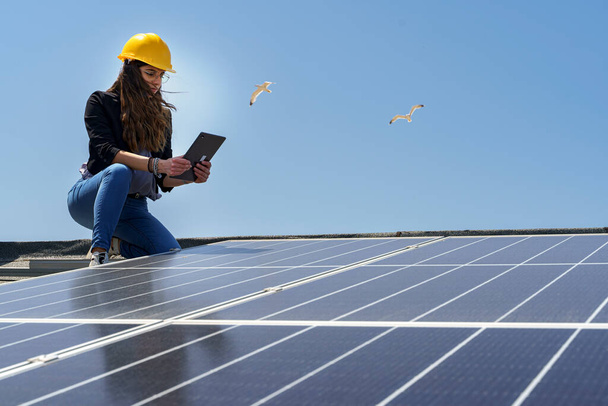 Vrouwelijke ingenieur in een gele harde hoed controleert de conditie van een fotovoltaïsch systeem op het dak van een huis met een tablet in haar hand - Foto, afbeelding