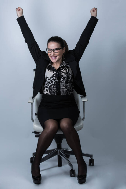 White business woman with collected hair and eyeglasses, dressed in black suit and patterned shirt, rejoices in victory sitting in her armchair, isolated on white background - Φωτογραφία, εικόνα