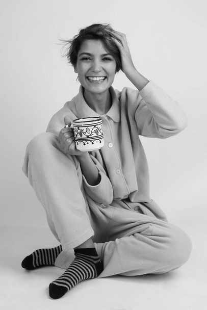 girl with bob hair and pajamas holds a cup sitting on the floor, isolated on light background, black and white - Photo, Image