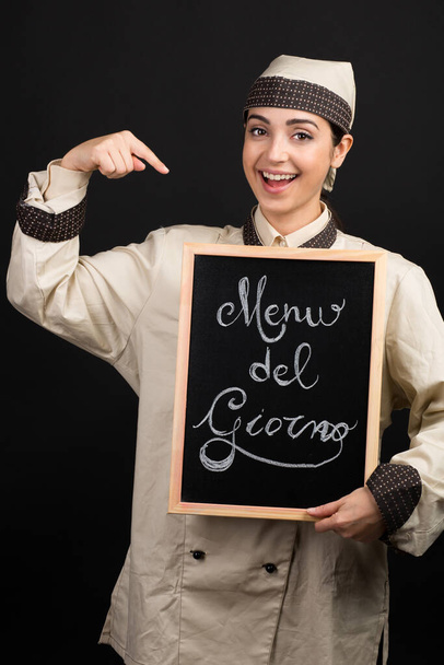Glimlachende chef-kok in uniform houdt een bord in zijn hand met daarop geschreven in het Italiaans "Menu del giorno" wat "Menu van de dag" betekent, geïsoleerd op oranje achtergrond - Foto, afbeelding