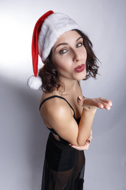 Beautiful brunette girl in a black dress and santa hat is dancing and having fun, isolated on white background. - 写真・画像