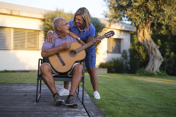 ώριμος άντρας παίζει κιθάρα ενώ κάθεται με τη γυναίκα του δίπλα στο σπίτι - Φωτογραφία, εικόνα