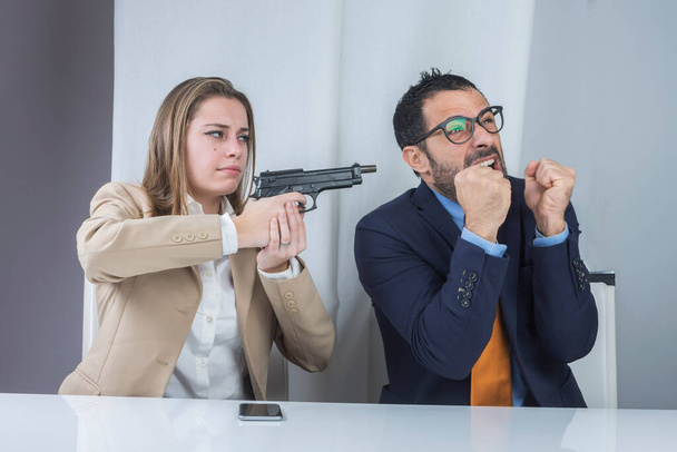 Kaksi kollegaa istuu työpaikalla, vihainen sihteeri osoittaa aseella epätoivoista pomoaan. - Valokuva, kuva