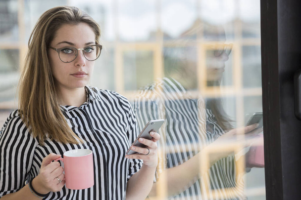 Fair-skinned blonde meisje in bril en een gestreept shirt staat te kijken naar haar smartphone en het houden van een kopje koffie, bij het raam van haar kantoor - Foto, afbeelding