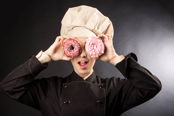 Hymyilevä kokki soittaa kauniisti värillisillä donitseilla käyttäen niitä kaukoputkina, eristettynä mustalla taustalla. - Valokuva, kuva
