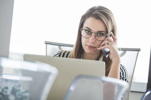 Młoda blond pracownik biurowy w pasiastej koszuli siedzi przy biurku i rozmawia przez telefon przed komputerem - Zdjęcie, obraz