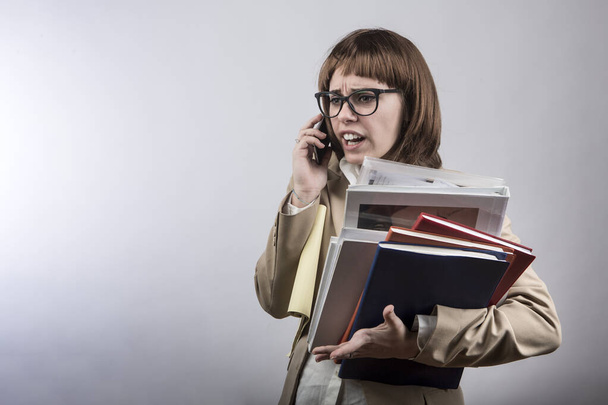 girl with brown bob hair, glasses and dress is holding a folder of documents in one hand and using her smartphone with the other with a very angry face, isolated on a white background - Foto, Imagem