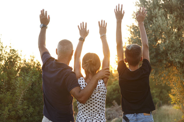 Сім'я з трьох людей, ззаду, обіймає один одного як знак союзу, що стоїть перед заходом сонця в природному контексті - Фото, зображення