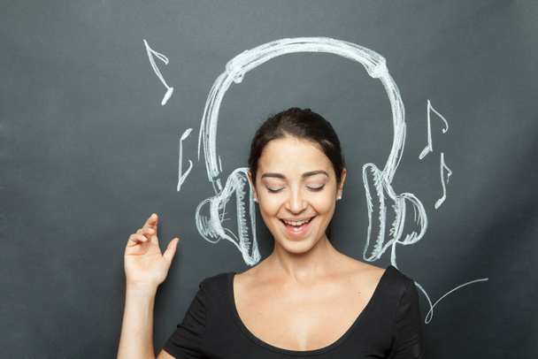 Dark-haired girl pretends to listen to music thanks to headphones designed on the dark background - Foto, Bild