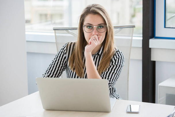 Блондинка менеджер в очках и в полосатой рубашке работает в офисе сидя перед ее рабочим местом - Фото, изображение