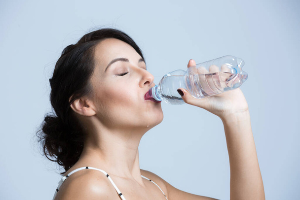 Fehér nő fekete hajjal, fehér csíkos kezeslábasban, egy kis üveg vízből iszik, fehér alapon elszigetelve. - Fotó, kép