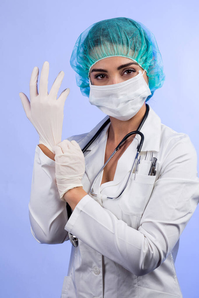 Female doctor in white coat, white face mask, green bonnet for hair is intent on putting on latex gloves, isolated on white background - Foto, afbeelding