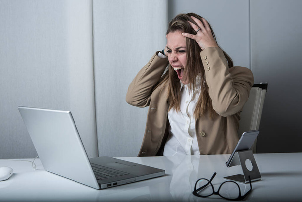 manager woman in jacket and shirt gets desperate and brings her hands to her head sitting in front of her laptop - 写真・画像