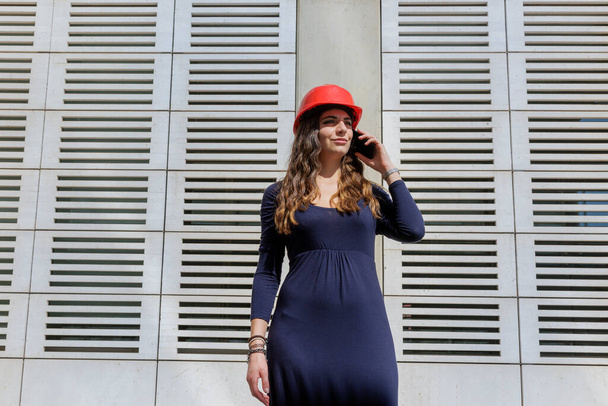 Elegantly dressed woman in red hard hat speaks with serious expression on the phone in modern urban setting - Foto, Imagem