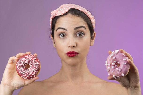 schönes Mädchen mit weißem, schwarzem Haar und hellem Hemd, spielt und hat Spaß mit 2 farbigen Donuts, isoliert auf rosa Hintergrund - Foto, Bild