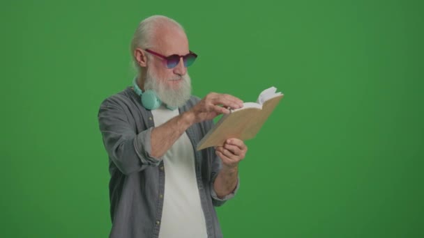 Tela Verde. Retrato de um velho inteligente com barba cinza, óculos engraçados e fones de ouvido, lendo um livro. Um Idoso Vira-se Através das Páginas de um Livro. Lazer, Hobby Concept. - Filmagem, Vídeo