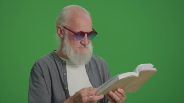 Zöld vászon. Portré egy okos öregemberről, szürke szakállal és vicces szemüveggel, aki könyvet olvas. Egy Idős ember átlapozza egy könyv oldalait. Szabadidő, Hobby koncepció. - Felvétel, videó
