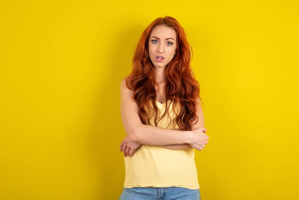 Shocked embarrassed young red-haired wearing yellow t-shirt yellow studio background keeps mouth widely opened. Hears unbelievable novelty stares in stupor - 写真・画像